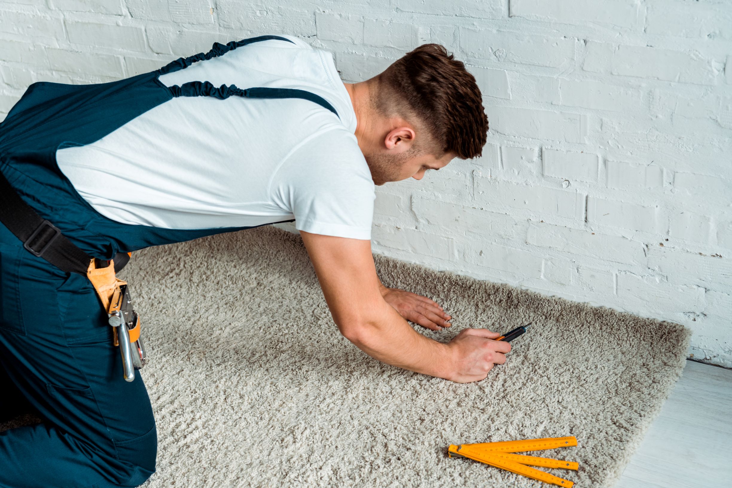 A worker in blue overalls kneeling while using a utility knife to trim carpet along a white brick wall.